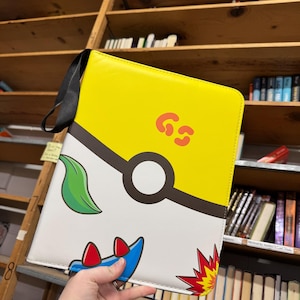 May include: A rectangular yellow and white binder featuring a black circular graphic resembling a ball, a green leaf illustration, a blue shape with red accents, and a red starburst design. The letters GS are printed in orange on the yellow section.