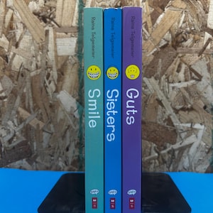 May include: Three graphic novels by Raina Telgemeier titled Smile, Sisters, and Guts, standing upright between two black metal bookends. The spines are light green, blue, and purple, each featuring a yellow smiley face icon and the Graphix logo.