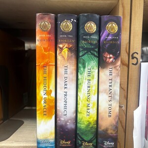 May include: Four books from the Riordan series, displayed on a wooden shelf. The spines feature titles like "The Hidden Oracle" and "The Tyrant's Tomb," with the Disney Hyperion logo. Each book has a unique color scheme, including orange, purple, and green.