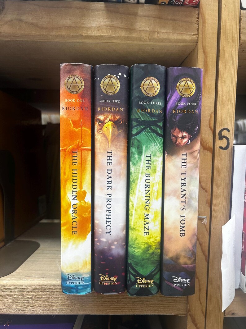 May include: Four books from the Riordan series, displayed on a wooden shelf. The spines feature titles like "The Hidden Oracle" and "The Tyrant's Tomb," with the Disney Hyperion logo. Each book has a unique color scheme, including orange, purple, and green.
