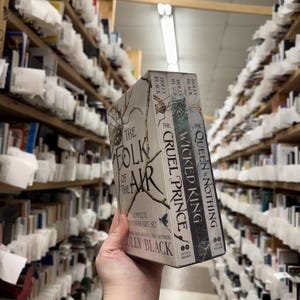 May include: A complete paperback gift set of "The Folk of the Air" series by Holly Black. The set includes "The Folk of the Air", "The Cruel Prince", and "The Wicked King & Queen Nothing" books. The books are held in a bookstore.