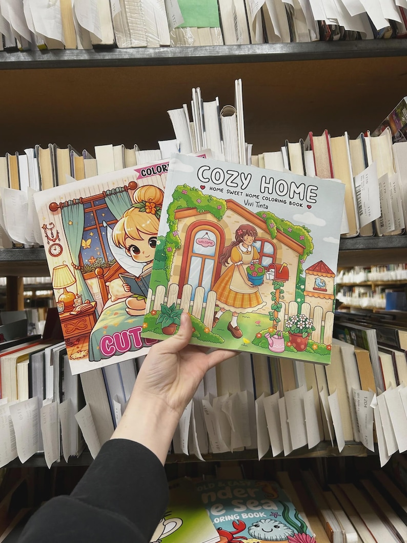 May include: Two coloring books are held up in front of a library shelf. The book on the right is titled "COZY HOME" with a picture of a house and a woman. The book on the left has the word "COLOR" and a picture of a girl.