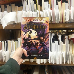 May include: A hardcover book titled "The Trials of Apollo: The Tyrant's Tomb" by Rick Riordan. The cover features a purple and gold design with illustrations of characters in action. The book is held in front of a bookshelf filled with other books.