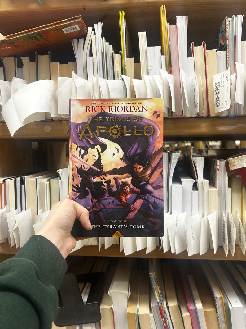 May include: A hardcover book titled "The Trials of Apollo: The Tyrant's Tomb" by Rick Riordan. The cover features a purple and gold design with illustrations of characters in action. The book is held in front of a bookshelf filled with other books.