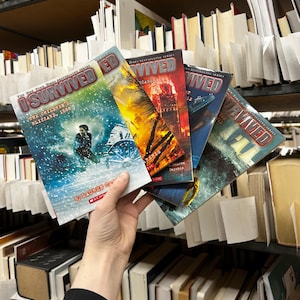 May include: A hand holding a stack of "I Survived" books from the New York Times Bestselling Series. The covers feature illustrations of historical events, including a blizzard, a fire, and a ship. The books are in a library setting.