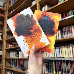 May include: Two paperback books titled "Spark of the Everflame" and "Everflame" are held in front of a bookshelf. The covers feature a silhouette and fiery orange and yellow backgrounds. The author's name, Cole, is visible.