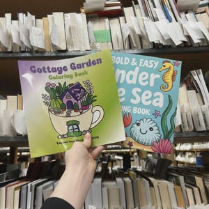 May include: Two coloring books are held up in front of a bookshelf. The first book is titled "Cottage Garden Coloring Book" and features a teacup with a house and plants. The second book is titled "Bold & Easy Under the Sea Coloring Book" with a seahorse and sea creatures.