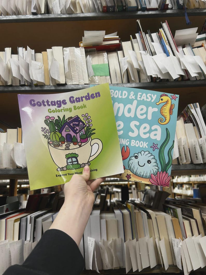 May include: Two coloring books are held up in front of a bookshelf. The first book is titled "Cottage Garden Coloring Book" and features a teacup with a house and plants. The second book is titled "Bold & Easy Under the Sea Coloring Book" with a seahorse and sea creatures.