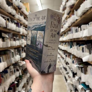 May include: A boxed set of "The Folk of the Air" complete paperback gift set. The box is gray with the book titles and cover art visible. The titles include "The Cruel Prince", "The Wicked King", and "The Queen of Nothing". The background shows bookshelves.
