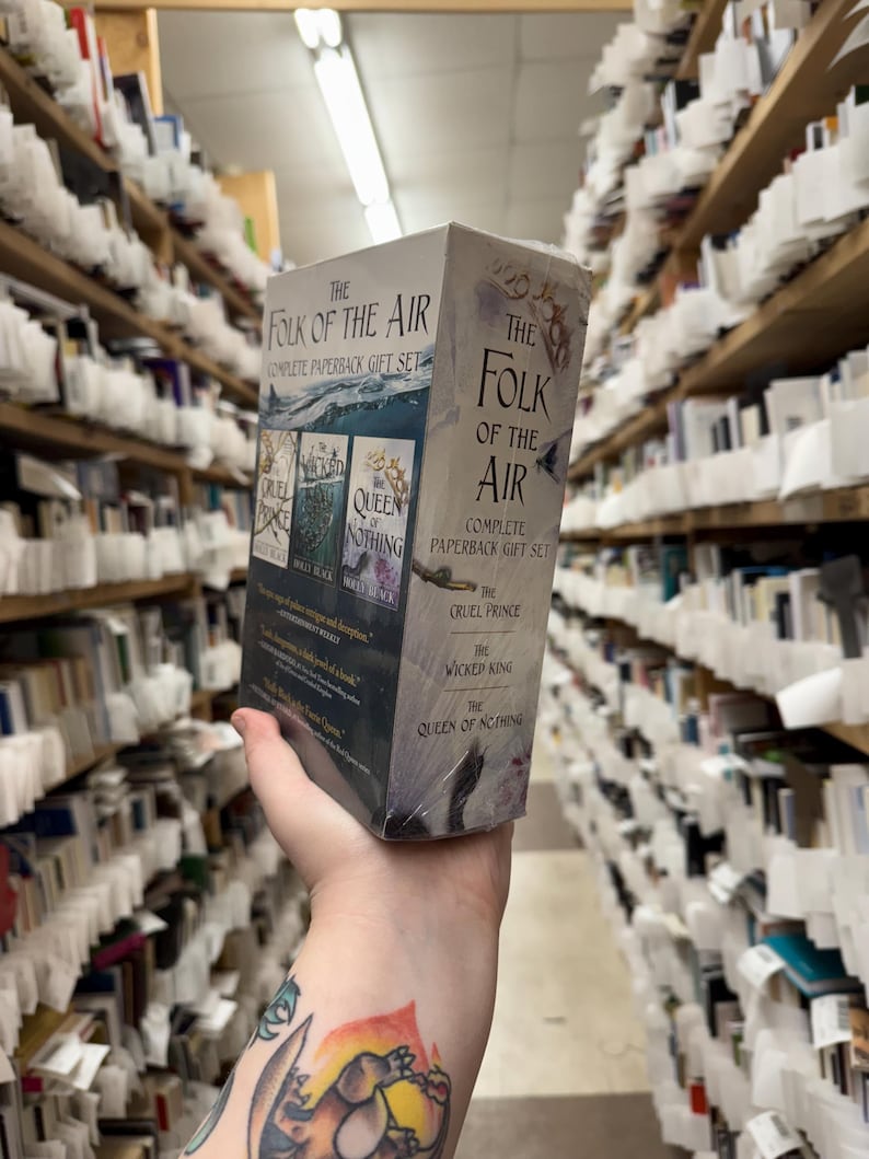 May include: A boxed set of "The Folk of the Air" complete paperback gift set. The box is gray with the book titles and cover art visible. The titles include "The Cruel Prince", "The Wicked King", and "The Queen of Nothing". The background shows bookshelves.