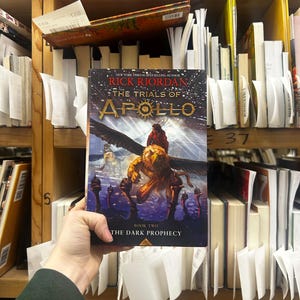 May include: A hardcover book titled "The Trials of Apollo: The Dark Prophecy" by Rick Riordan. The cover features a winged creature and a figure riding it, set against a dark background. The book is held in front of a bookshelf.