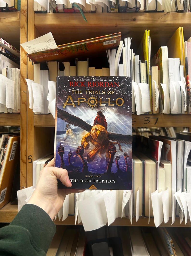 May include: A hardcover book titled "The Trials of Apollo: The Dark Prophecy" by Rick Riordan. The cover features a winged creature and a figure riding it, set against a dark background. The book is held in front of a bookshelf.