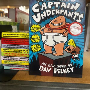 May include: A hardcover book titled "The Adventures of Captain Underpants" by Dav Pilkey. The cover features a cartoon character in underwear, a cape, and a red scarf. The book is on a shelf with other books.