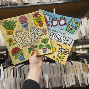 May include: Two coloring books are held up in front of a bookshelf. The first book is yellow with the text "BOLD AND EASY FLOWERS & PLANTS COLORING BOOK". The second book is blue with the text "SUMMER COLORING BOOK".