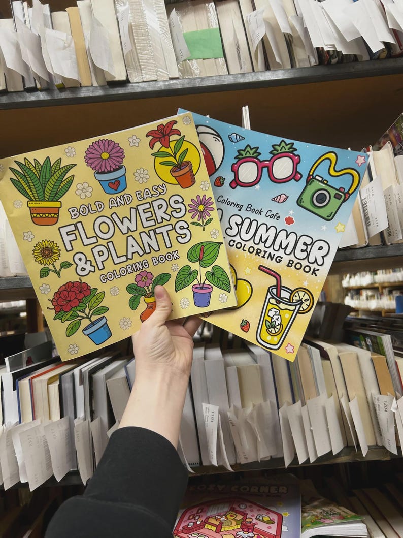 May include: Two coloring books are held up in front of a bookshelf. The first book is yellow with the text "BOLD AND EASY FLOWERS & PLANTS COLORING BOOK". The second book is blue with the text "SUMMER COLORING BOOK".