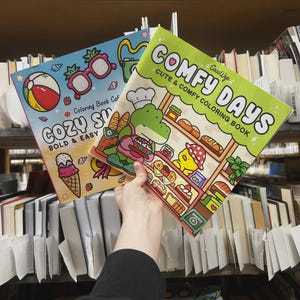 May include: Two coloring books are held up in front of a bookshelf. One book is titled "COZY SUMMER" with a beach ball and ice cream cone illustration. The other book is titled "COMFY DAYS" with a bakery-themed illustration. Both books are labeled "Coloring Book Caf".