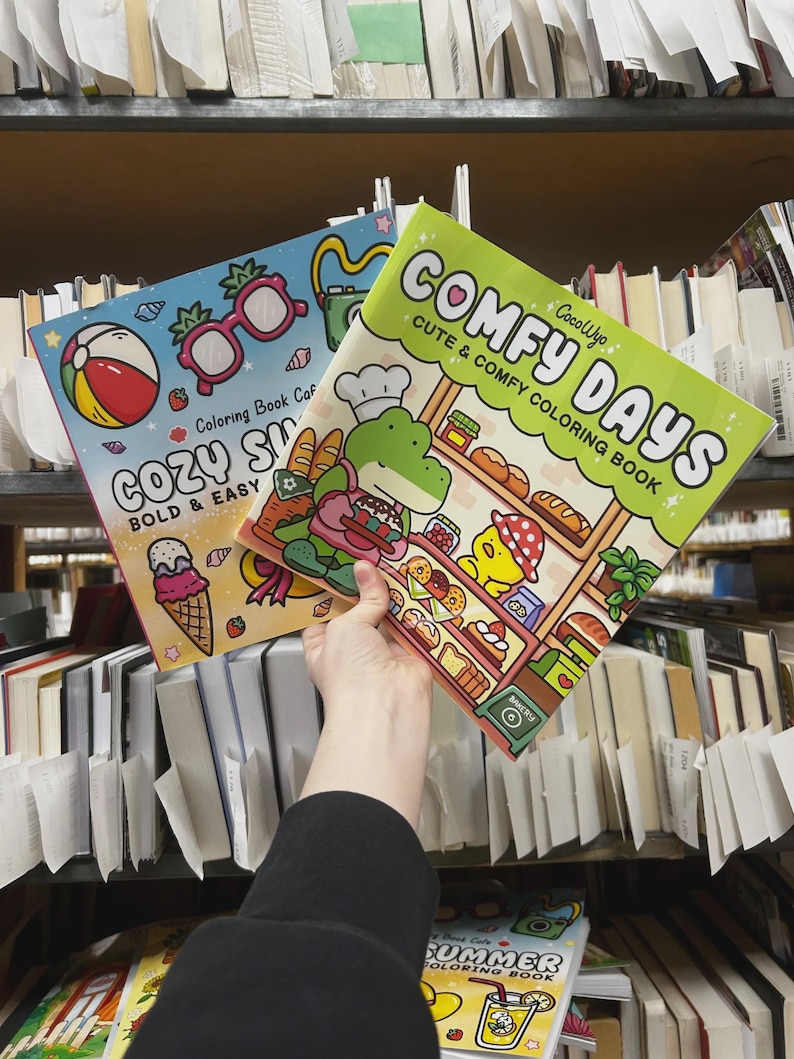 May include: Two coloring books are held up in front of a bookshelf. One book is titled "COZY SUMMER" with a beach ball and ice cream cone illustration. The other book is titled "COMFY DAYS" with a bakery-themed illustration. Both books are labeled "Coloring Book Caf".