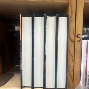 May include: Four hardcover books with white pages and colorful spines are arranged on a wooden shelf. The spines have a dark blue or purple color with abstract designs. The books are standing upright, with the pages facing forward.