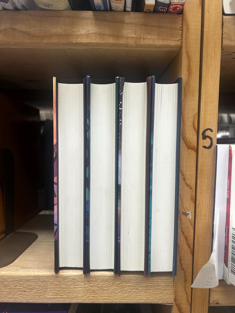 May include: Four hardcover books with white pages and colorful spines are arranged on a wooden shelf. The spines have a dark blue or purple color with abstract designs. The books are standing upright, with the pages facing forward.