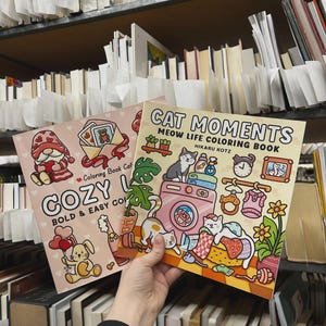 May include: Two coloring books are held up in front of a bookshelf. One book is titled "COZY" with a gnome and bunny illustration. The other book is titled "CAT MOMENTS" with a cat-themed illustration. The books are in a library setting.