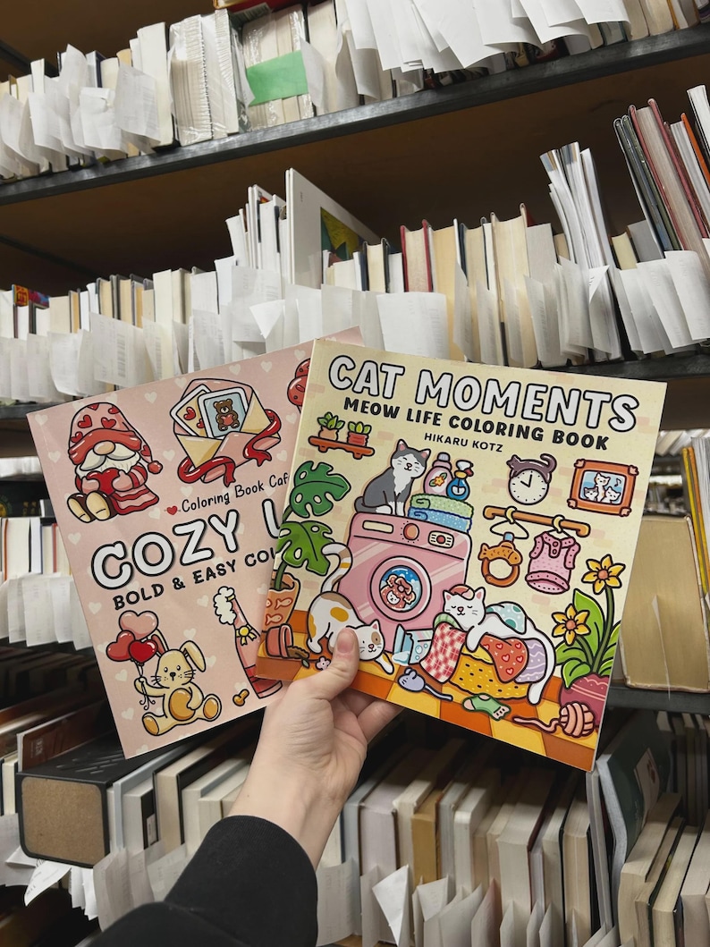 May include: Two coloring books are held up in front of a bookshelf. One book is titled "COZY" with a gnome and bunny illustration. The other book is titled "CAT MOMENTS" with a cat-themed illustration. The books are in a library setting.