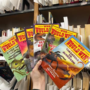 May include: A hand holds a stack of children's books from the "Who Would Win?" series. The covers feature illustrations of animals and creatures, including a tarantula, scorpion, dinosaur, and ant. The books are surrounded by other books on shelves.