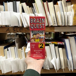 May include: A red Brain Quest Reading Basics box for Grade 2, ages 7-8, with the text "56 Stories with Questions and Answers." The box is held in front of a shelf of books in a library or bookstore.