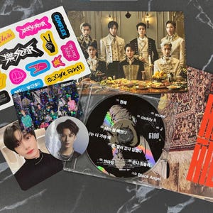 May include: A collection of K-pop merchandise, including a CD, photo cards, stickers, and a poster. The CD is black with white text and a silver reflective surface. The stickers are colorful with various designs and text. The poster has red text.