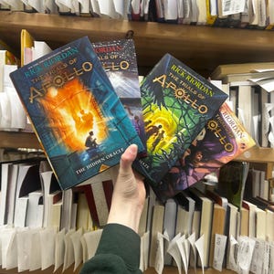 May include: Several copies of "The Trials of Apollo" by Rick Riordan are held in front of a bookshelf. The book covers feature vibrant illustrations with titles like "The Hidden Oracle" and "The Burning Maze". The books are surrounded by other books and shelf labels.