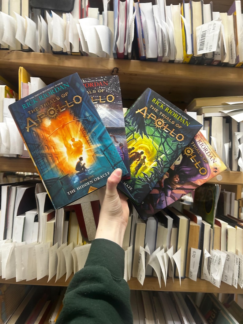 May include: Several copies of "The Trials of Apollo" by Rick Riordan are held in front of a bookshelf. The book covers feature vibrant illustrations with titles like "The Hidden Oracle" and "The Burning Maze". The books are surrounded by other books and shelf labels.