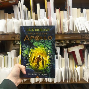 May include: A hardcover book titled "The Trials of Apollo: The Burning Maze" by Rick Riordan. The cover features a green and yellow maze with three running figures. The book is held in front of a shelf of other books.