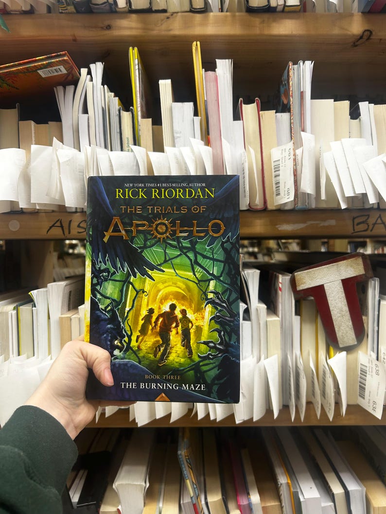 May include: A hardcover book titled "The Trials of Apollo: The Burning Maze" by Rick Riordan. The cover features a green and yellow maze with three running figures. The book is held in front of a shelf of other books.
