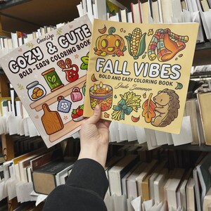 May include: Two coloring books titled "Cozy & Cute" and "Fall Vibes" are held in front of a bookshelf. The "Cozy & Cute" book features illustrations of cookies, tea, and other cozy items. The "Fall Vibes" book has a pumpkin, corn, and hedgehog illustration.
