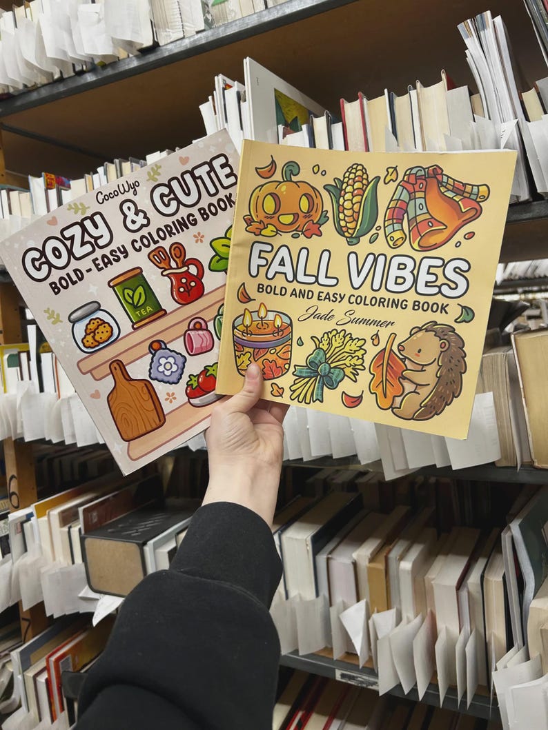 May include: Two coloring books titled "Cozy & Cute" and "Fall Vibes" are held in front of a bookshelf. The "Cozy & Cute" book features illustrations of cookies, tea, and other cozy items. The "Fall Vibes" book has a pumpkin, corn, and hedgehog illustration.