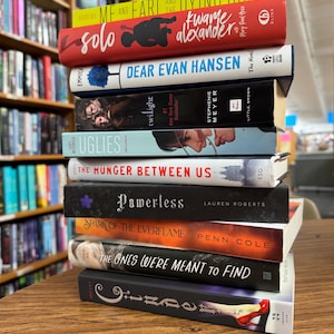 May include: A stack of hardcover books with various titles and authors. The books have colorful covers and include titles like "Me and Earl and the Dying Girl," "Solo," "Dear Evan Hansen," and "Twilight." The books are stacked on a wooden surface, with a bookshelf in the background.