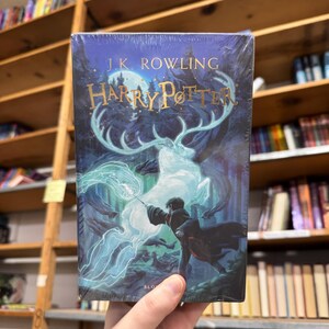 May include: A hardcover book titled "Harry Potter" by J.K. Rowling. The cover features a dark blue background with a depiction of a wizard casting a spell, with a glowing stag and forest imagery. The book is held in a person's hand.