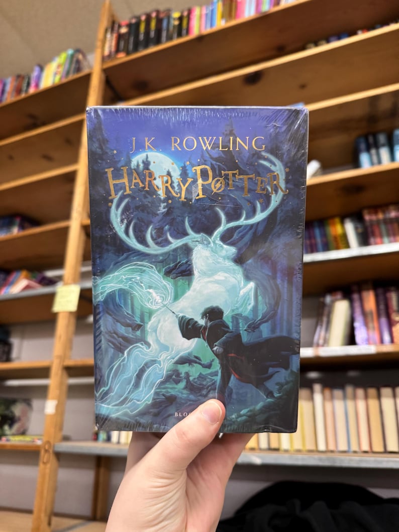 May include: A hardcover book titled "Harry Potter" by J.K. Rowling. The cover features a dark blue background with a depiction of a wizard casting a spell, with a glowing stag and forest imagery. The book is held in a person's hand.