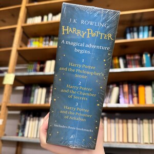 May include: A boxed set of the first three Harry Potter books by J.K. Rowling. The covers are dark blue with gold lettering and include the titles "Harry Potter and the Philosopher's Stone", "Harry Potter and the Chamber of Secrets", and "Harry Potter and the Prisoner of Azkaban".