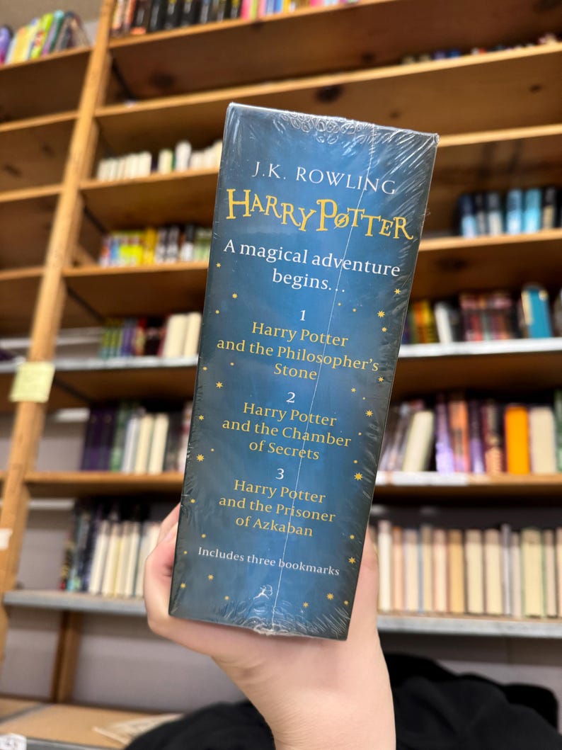May include: A boxed set of the first three Harry Potter books by J.K. Rowling. The covers are dark blue with gold lettering and include the titles "Harry Potter and the Philosopher's Stone", "Harry Potter and the Chamber of Secrets", and "Harry Potter and the Prisoner of Azkaban".