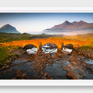 Könnte beinhalten: Eine Steinbrücke mit drei Bögen überspannt einen felsigen Bach in einer schottischen Hochlandlandschaft. Die Berge im Hintergrund sind in Nebel gehüllt und der Himmel ist zartblau.