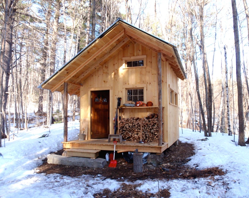 May include: A small wooden cabin with a porch and a woodpile in front of it. The cabin is surrounded by snow-covered trees.