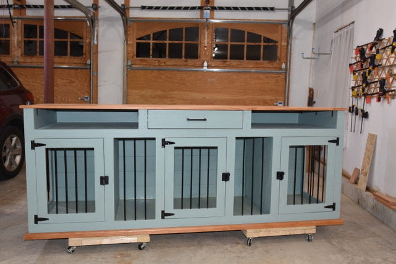 Op de afbeelding: Een op maat gemaakte hondenbench met een lichtblauwe verf en een bovenkant van natuurlijk hout. De bench heeft drie aparte compartimenten met zwarte metalen staven en deuren, en een lade. Gemonteerd op wielen, ongeveer 183 cm breed.