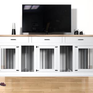 Op de afbeelding: Een witte en houten combinatie van hondenbench en tv-standaard. De unit heeft drie lades en drie hondenbench compartimenten met zwarte spijlen. Een zwarte tv staat erboven. Een hondenspeeltje ligt op de vloer.