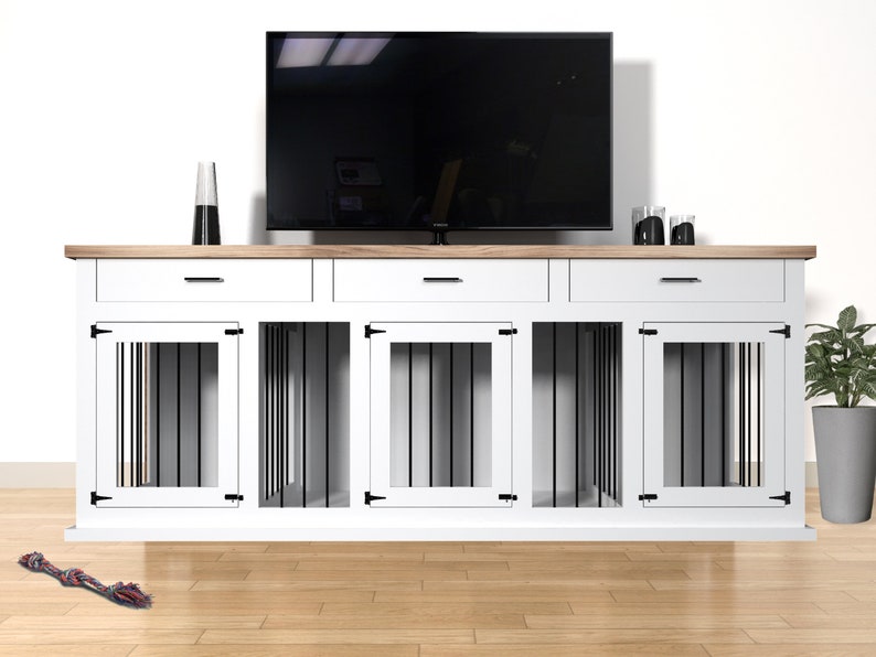 Op de afbeelding: Een witte en houten combinatie van hondenbench en tv-standaard. De unit heeft drie lades en drie hondenbench compartimenten met zwarte spijlen. Een zwarte tv staat erboven. Een hondenspeeltje ligt op de vloer.