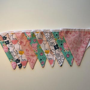 May include: A bunting with pink, green, and white triangular flags featuring a cartoon pig design. The flags are sewn together with white thread.