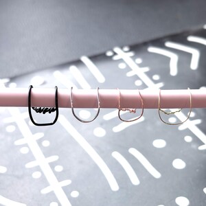 May include: Four minimalist earrings in black, silver, rose gold, and gold hang on a pink rod. Each earring features a unique, abstract design at the bottom, adding a touch of modern art to the jewelry.