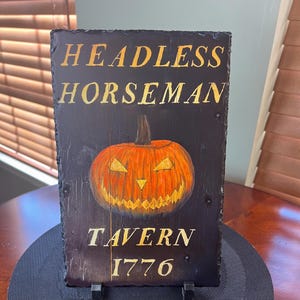 May include: A black slate sign with gold lettering that reads "Headless Horseman Tavern 1776". A hand-painted orange jack-o'-lantern with a menacing grin is featured in the centre of the sign.
