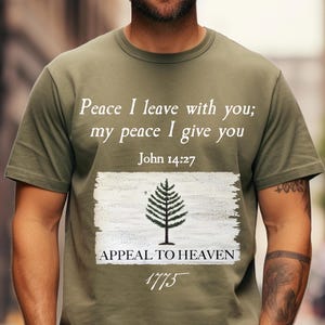 Könnte beinhalten: Olivengrünes T-Shirt mit einem weißen Flaggen-Design. Die Flagge zeigt einen Kiefernbaum und den Text "Appeal to Heaven 1775". Das Shirt trägt außerdem den Text "Peace I leave with you; my peace I give you John 14:27".