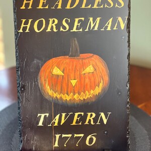 May include: A black slate sign with gold lettering that reads "Headless Horseman Tavern 1776". A carved wooden pumpkin with a menacing face is in the centre of the sign.