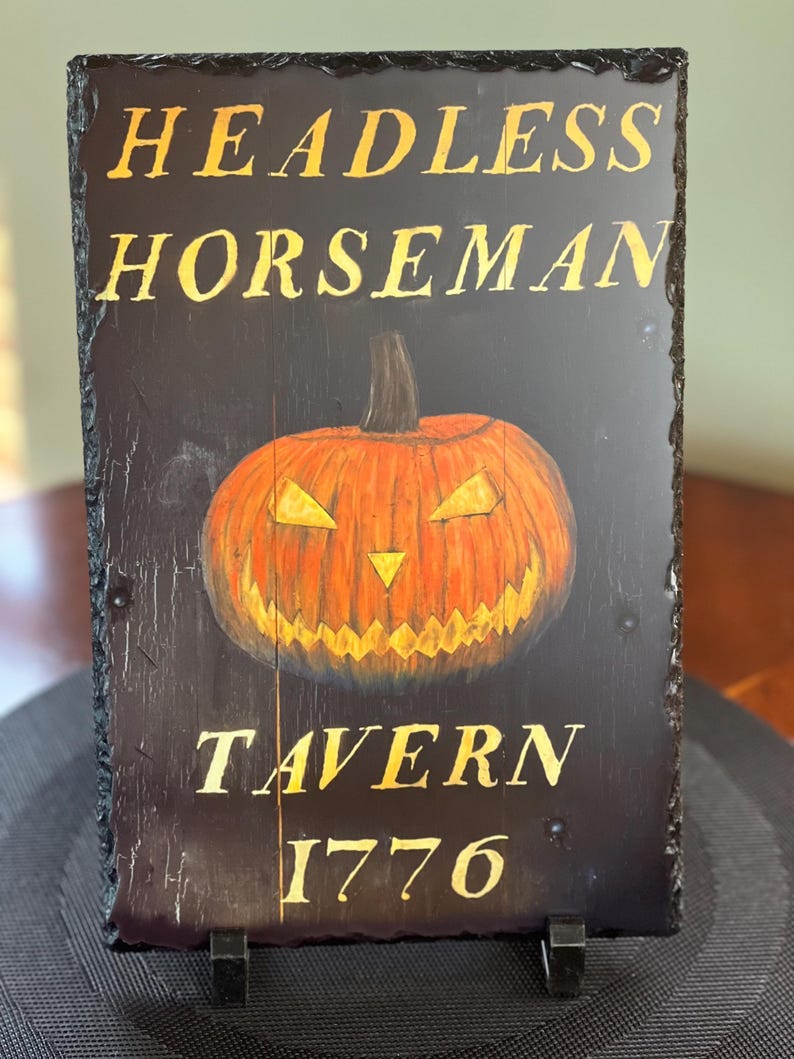 May include: A black slate sign with gold lettering that reads "Headless Horseman Tavern 1776". A carved wooden pumpkin with a menacing face is in the centre of the sign.